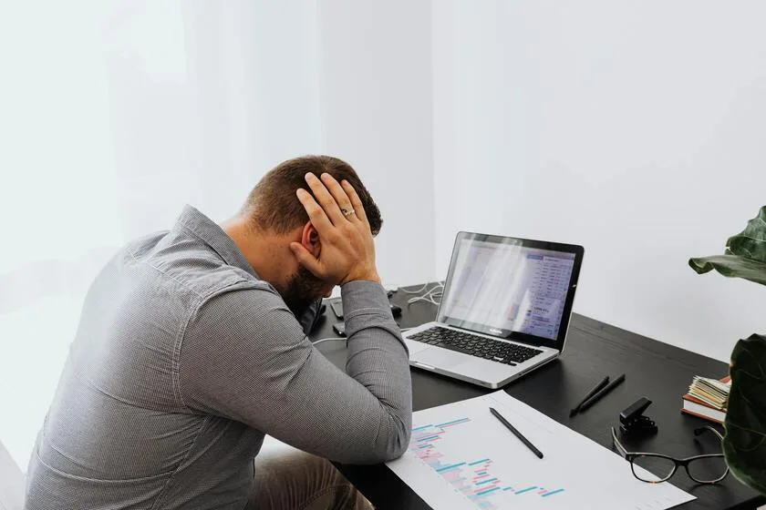 A man sits at a desk, visibly distressed, with his head resting in his hands, conveying a sense of frustration or overwhelm with his financial situation.