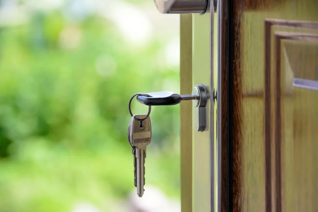 Close-up of a wooden door slightly ajar with a silver key inserted in the lock. Two additional keys on a keychain hang below. The background shows a blurred view of a green garden.