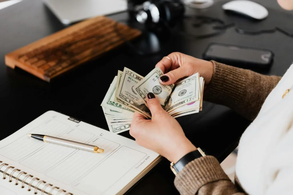 A person counts a stack of US dollar bills at a desk. Nearby are a notebook with a pen, a wooden keyboard, headphones, and a smartphone. The person wears a brown sweater and a black watch.