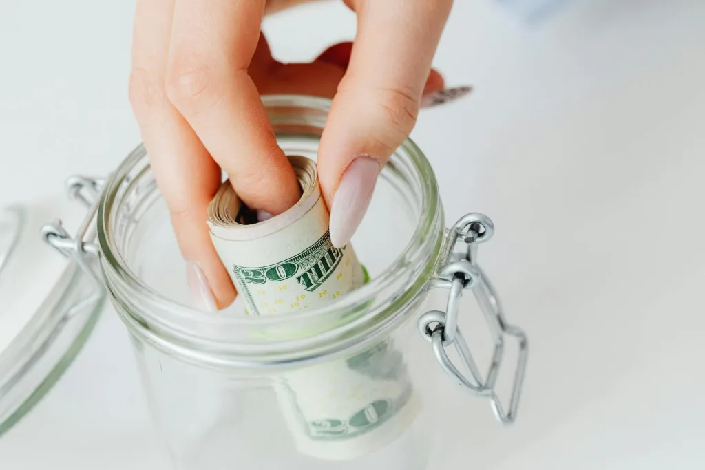 A person depositing coins into a jar with a lid, symbolizing saving and financial planning.